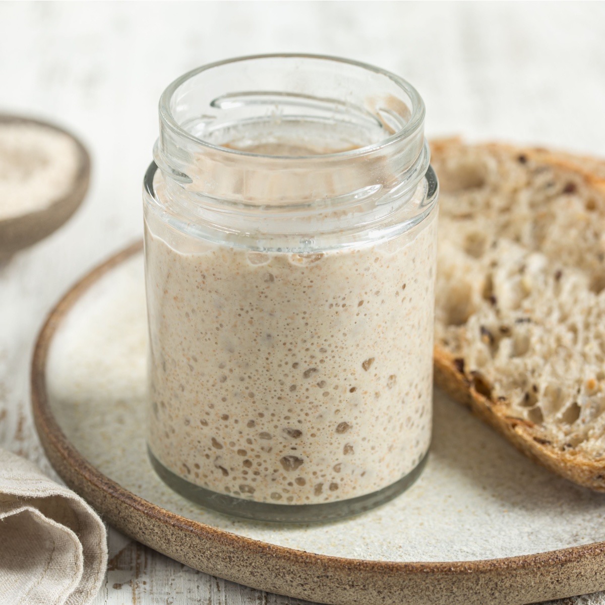 A jar of bubbly sourdough starter beside a slice of sourdough bread